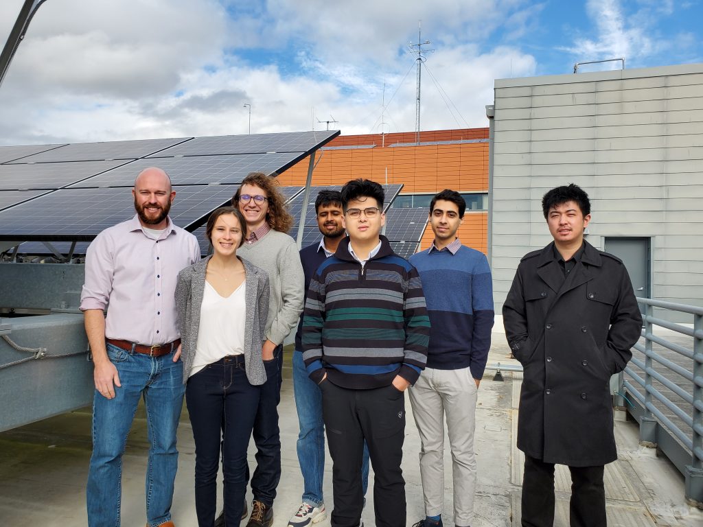 Graduate students and bald professor smile into the camera on a windy day while standing next to solar panels. 